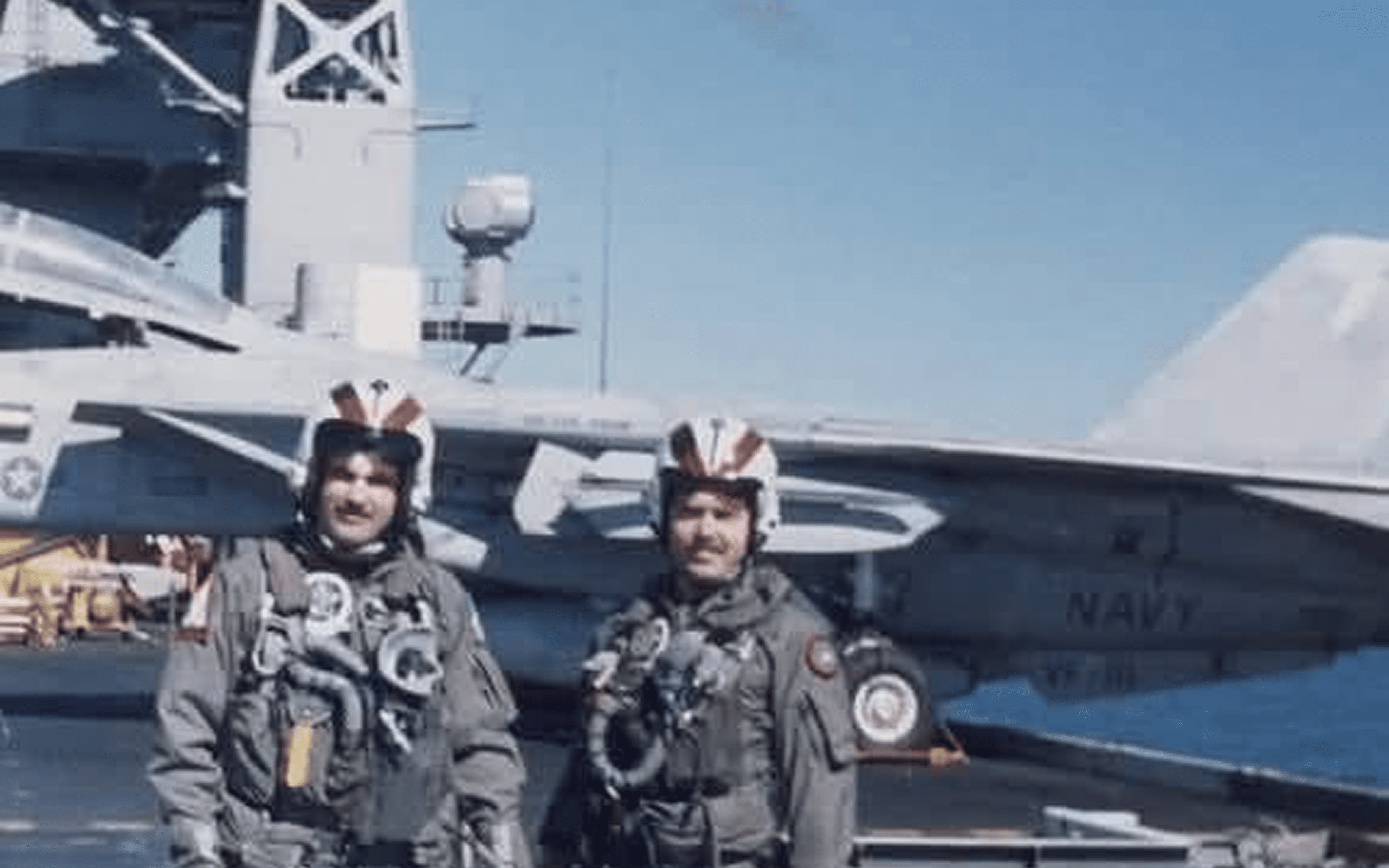 Two pilots in flight suits stand in front of a naval aircraft carrier.