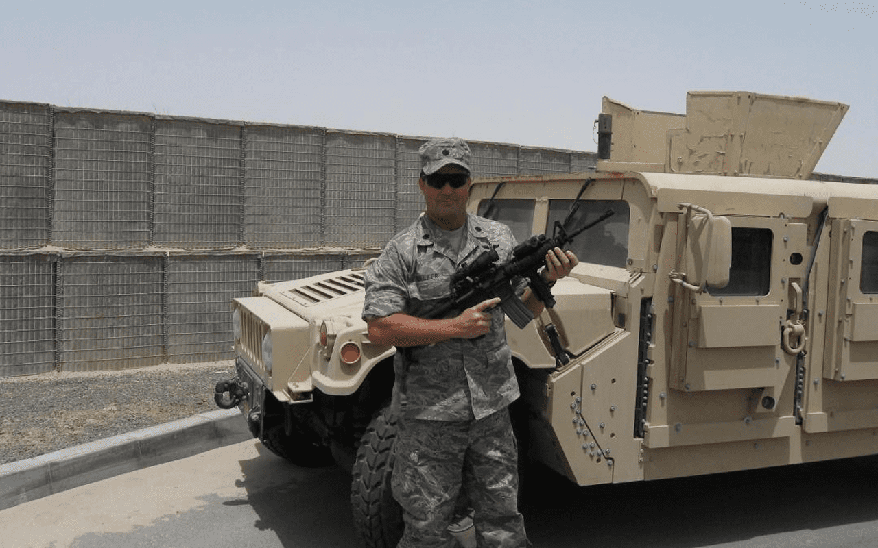 A soldier in camouflage stands with a rifle beside a military vehicle.