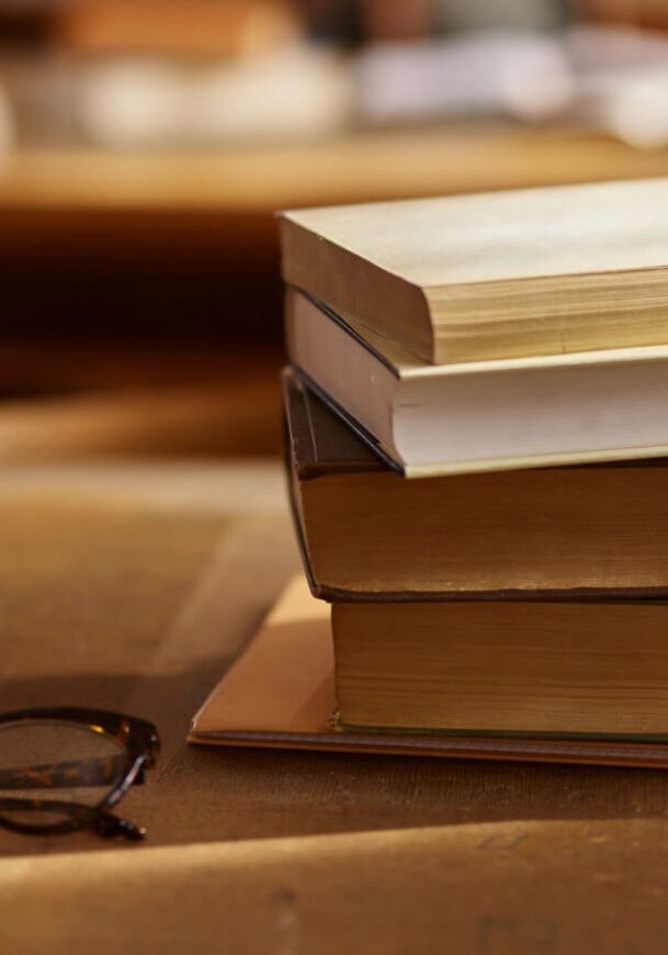 Stack of books and eyeglasses on a wooden table in warm light.